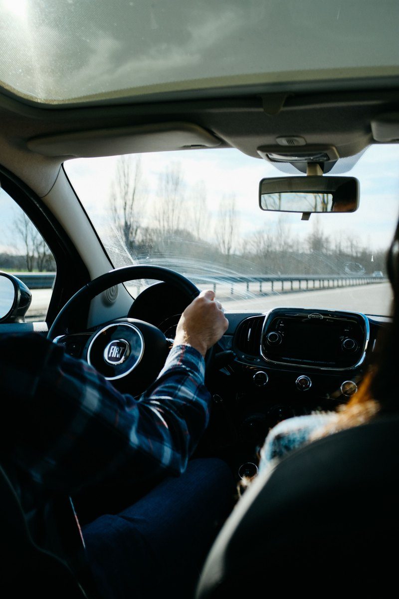 Driver in plaid shirt steering Fiat on highway with bare trees visible ahead